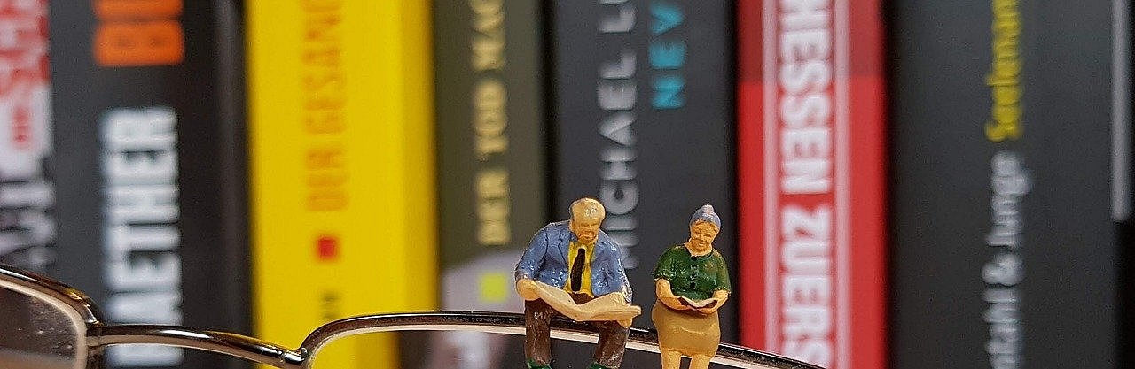 Ein älteres Paar (kleine Figuren) sitzt auf einem Brillenrand. Die Brille liegt auf einem geöffneten Buch.