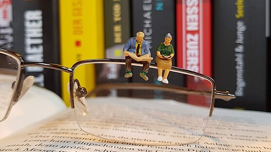 Ein älteres Paar (kleine Figuren) sitzt auf einem Brillenrand. Die Brille liegt auf einem geöffneten Buch.
